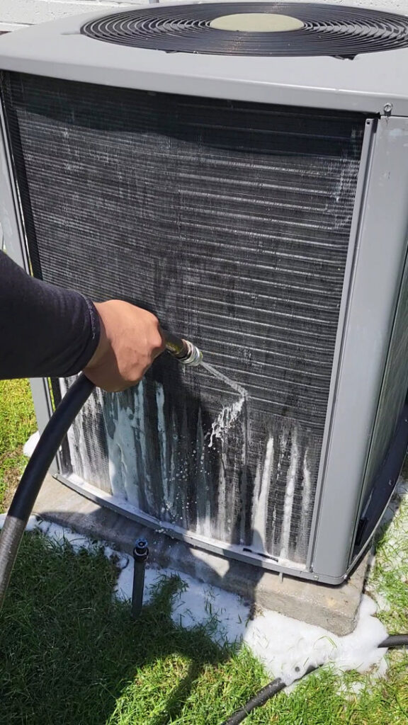 A technician cleaning the dirty coils of an outdoor HVAC unit with a hose for Modern Air Services in Fort Worth, TX.