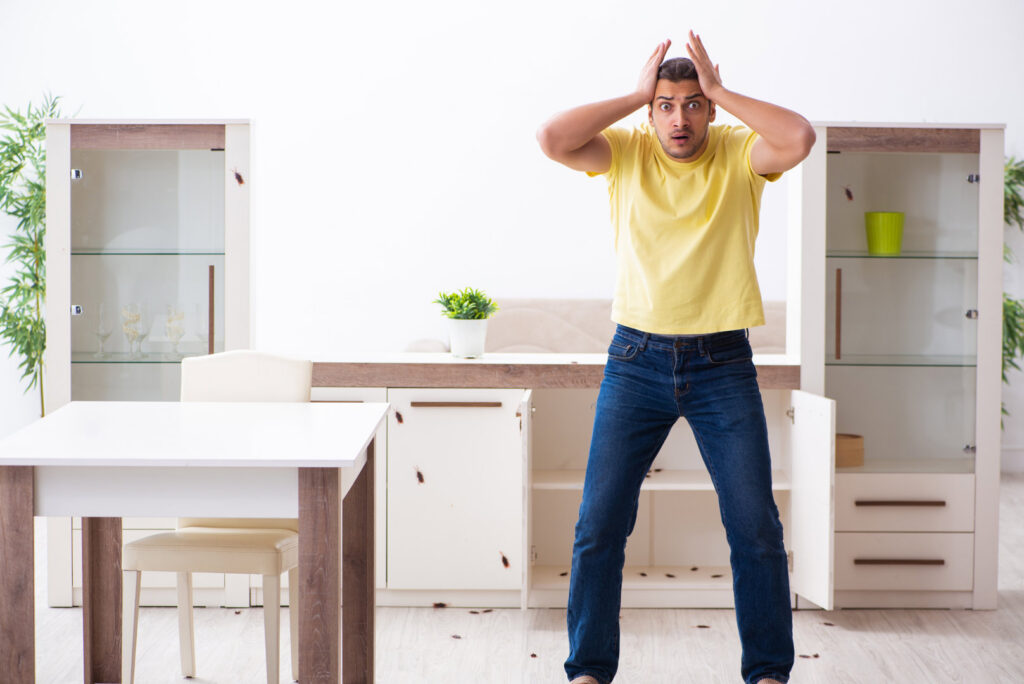 A man looking shocked at a severe cockroach infestation on the floor and furniture in a home, handled by Ares Pest Control in Biddeford, ME.