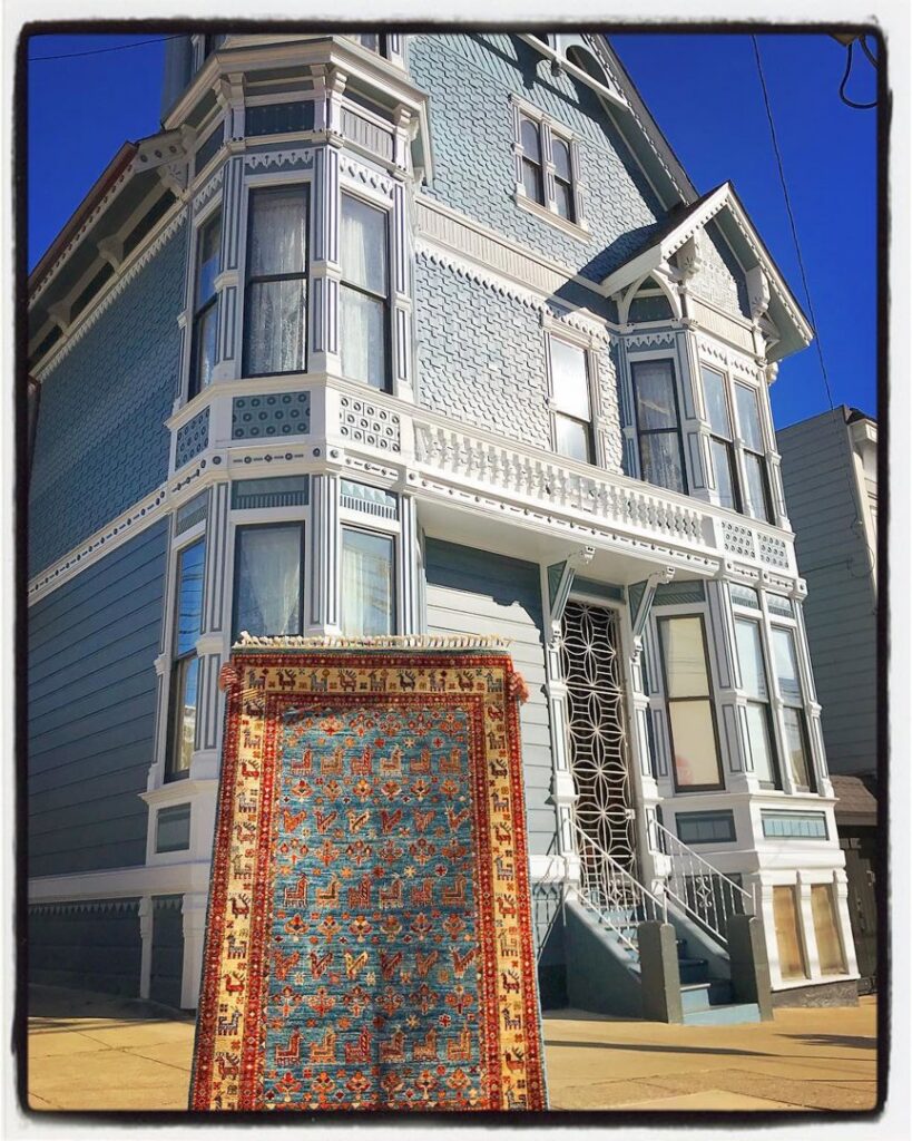 A vibrant patterned rug stands upright in front of a light blue Victorian house by Nomad Rugs in San Francisco, CA.