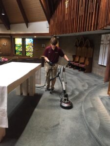 A technician from Yellow Finch Carpet Cleaning LLC using a rotary machine to clean blue carpet in a church, offering commercial services in Dover, NH.