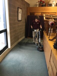 A technician from Yellow Finch Carpet Cleaning LLC using a rotary machine to clean blue carpet in a commercial hallway in Dover, NH.