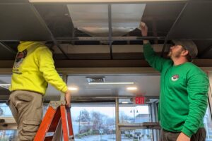 HVAC technicians installing or servicing a ceiling-mounted unit in a commercial building for WARM Global HVAC in East Providence, RI