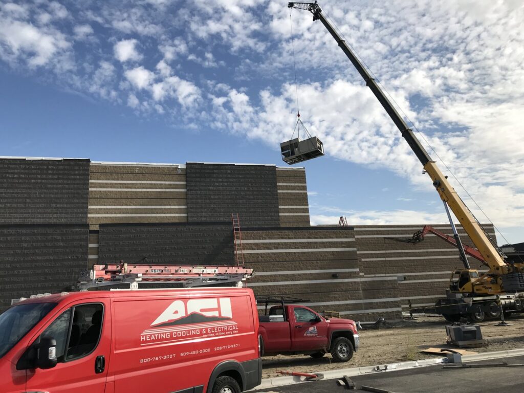 A commercial HVAC unit being lifted by a crane onto a building, with ACI Northwest Heating, Cooling & Electrical service vehicles on the job site in Dalton Gardens, ID.