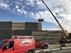 A commercial HVAC unit being lifted by a crane onto a building, with ACI Northwest Heating, Cooling & Electrical service vehicles on the job site in Dalton Gardens, ID.