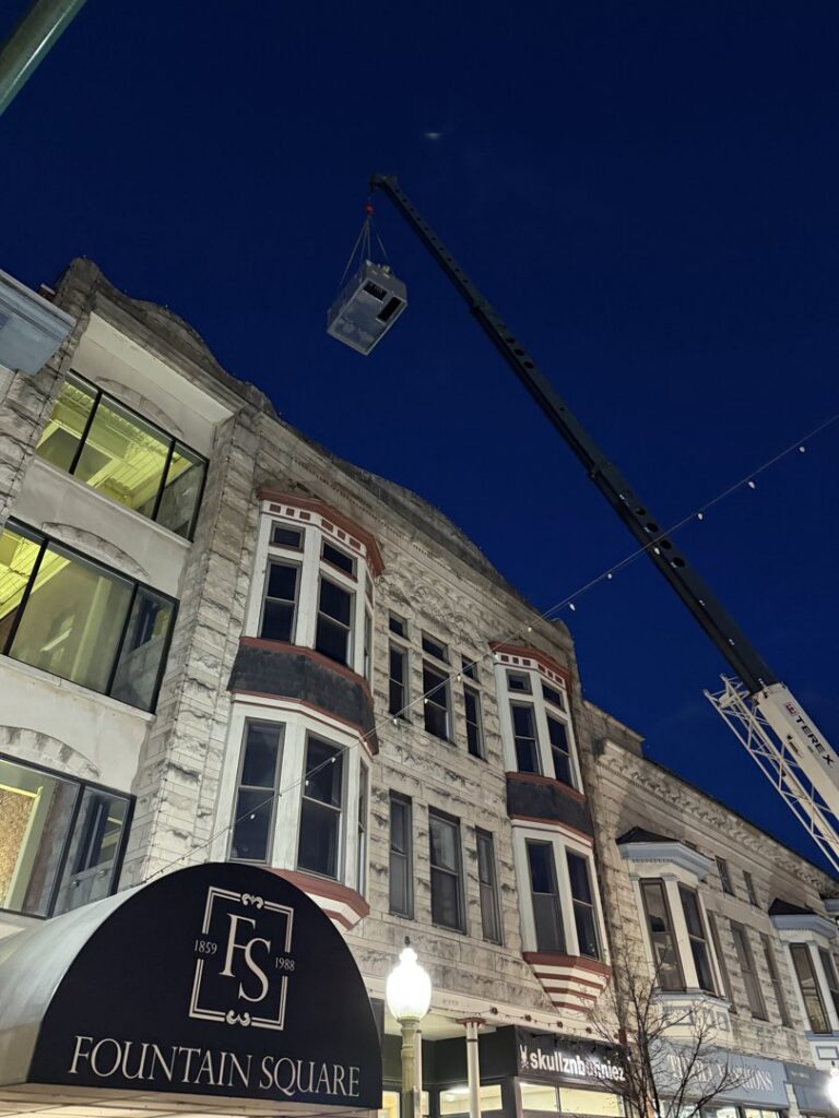 A crane lifting a commercial HVAC unit onto a building at night for Commercial Service in Bloomington, IN.