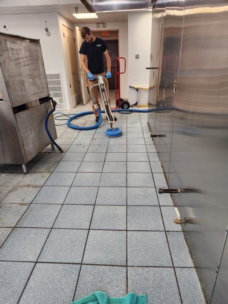 A technician cleaning a commercial kitchen tile floor with a rotary scrubber for LNC Steam Cleaning in Owasso, OK.