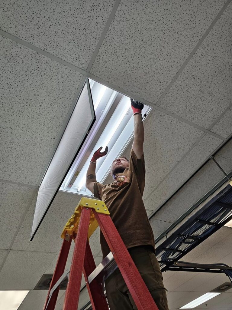 An electrician from Bee Thumb Electric in North Highlands, CA, repairing a commercial fluorescent light fixture on a ladder.