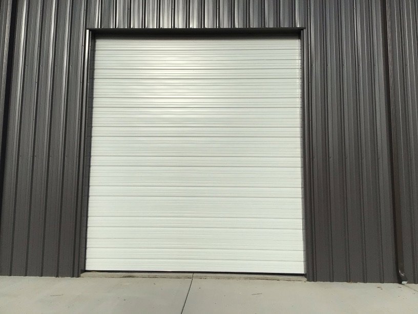 A white commercial roll-up garage door installed on a dark grey corrugated building by Magic City Door Huntsville in Birmingham, AL.