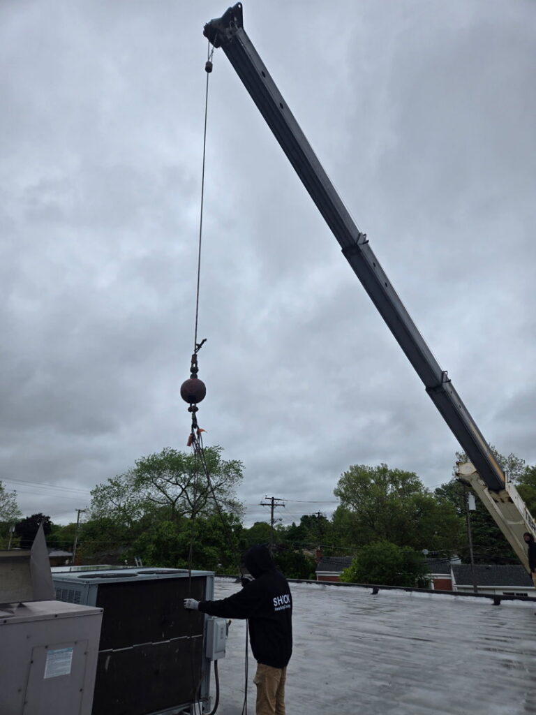 A worker on a commercial rooftop overseeing the installation of a large HVAC unit with a crane by Shook Heating And Cooling in Livonia, MI.