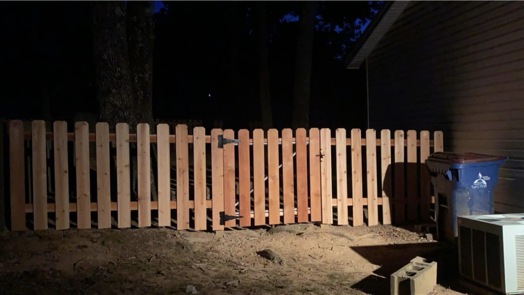 A completed wooden picket fence with a gate, installed by Fianna Fencing Company in Fort Smith, AR, illuminated at night.
