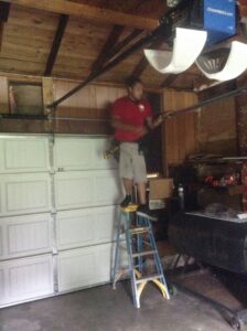 A contractor on a ladder working on a garage door opener mechanism, performing repairs for OklaHome Overhead Doors in Tulsa, OK