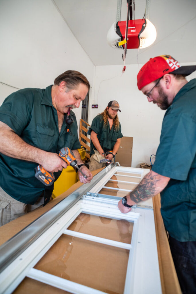 Two garage door contractors assembling a new garage door panel with windows using power tools for Annapolis Garage Doors Inc. in Annapolis, MD.