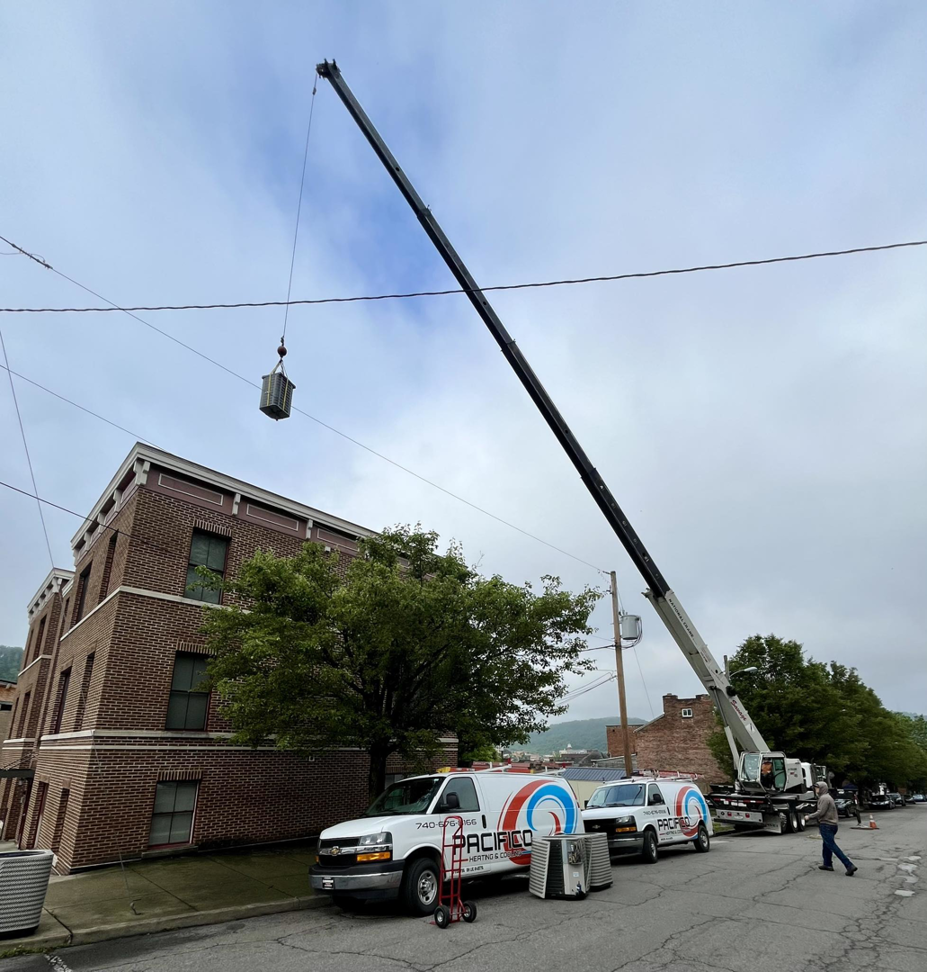 A crane lifting an HVAC unit with Pacifico Heating and Cooling service vans parked nearby in Shadyside, OH.