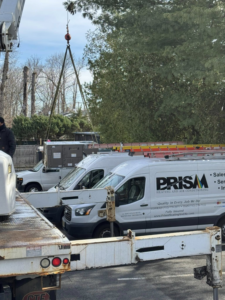A crane lifting a large commercial HVAC unit with Prism Heating & Air Conditioning vans on site in Toms River, NJ.
