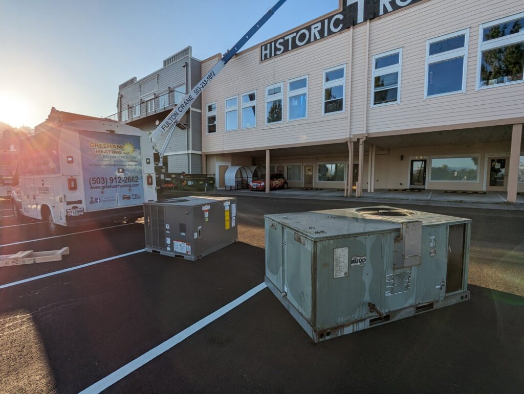 A crane lifting a new HVAC unit for installation, with an old unit nearby, by Gresham Heating and Air Conditioning in Wood Village, OR.