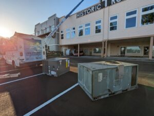 A crane lifting a new HVAC unit for installation, with an old unit nearby, by Gresham Heating and Air Conditioning in Wood Village, OR.