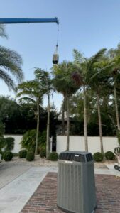 A crane operating to move HVAC equipment over palm trees, with an outdoor AC unit in the foreground by Air Zero in Largo, FL