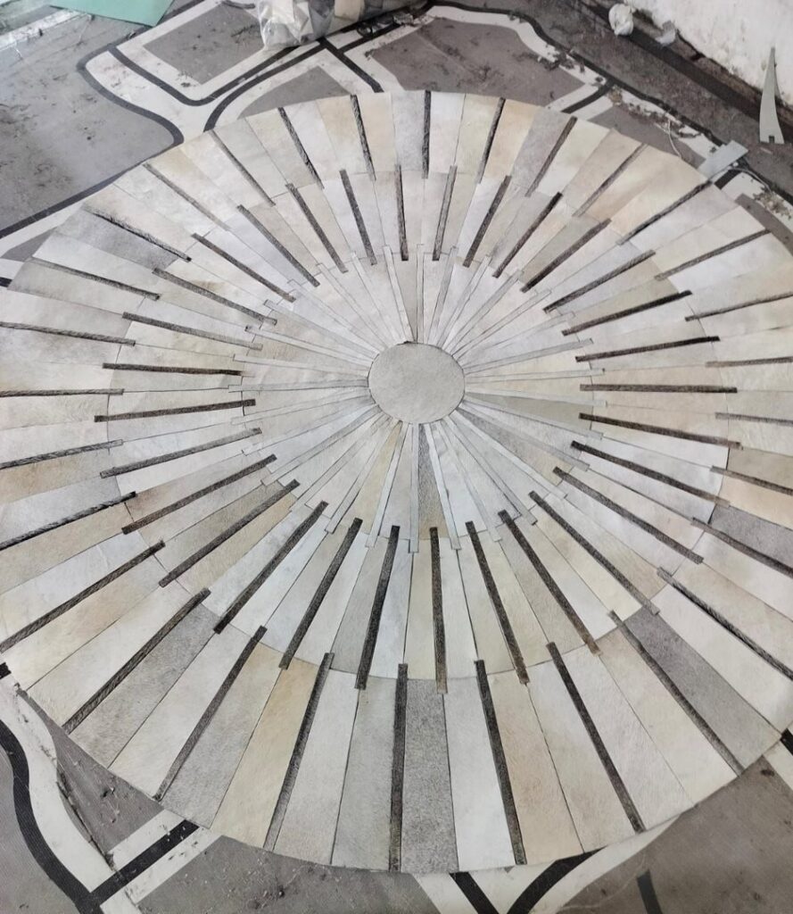 A custom circular rug made of light-colored strips being laid out on a floor, showcasing work by Makymo in Commerce, CA