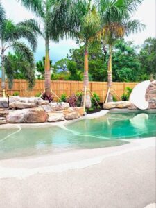A custom pool featuring a rock waterfall, slide, and surrounding palm trees, built by The Staycation Builders in Fort Wayne, IN.