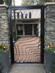 A custom black metal pedestrian gate with a unique wavy design and stone pillars installed by Craftsman Automatic Gates in Dallas, TX.