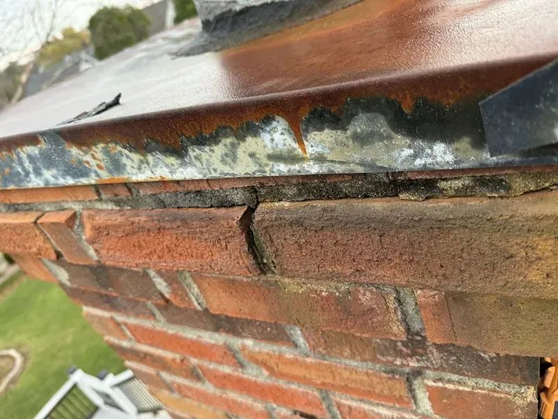 A close-up of a damaged chimney crown and flashing with a visible crack, showing repair needs by The Chimney Doctor in Louisville, KY.
