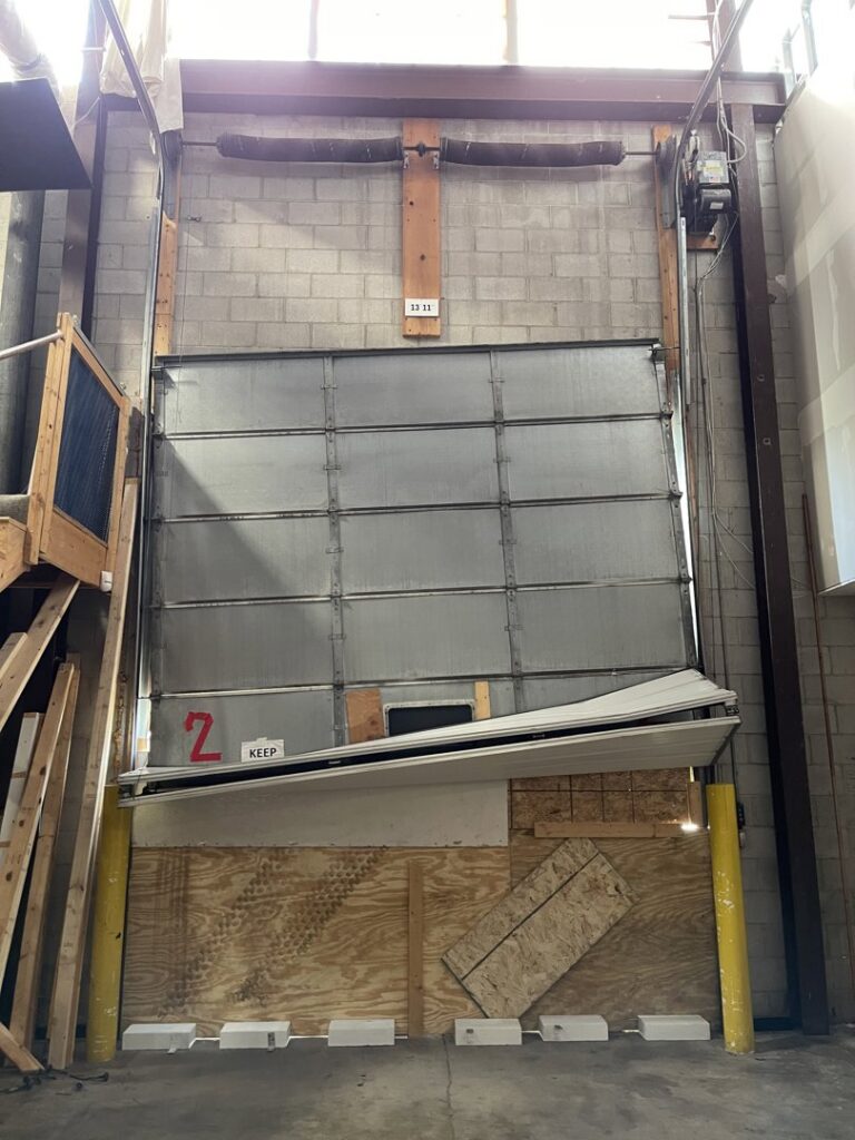 A severely damaged commercial garage door, bent and partially boarded up, requiring repair by Max Garage Door in Minneapolis, MN.