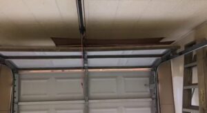 Interior view of a garage showing a white sectional door, opener rail, and damaged ceiling, indicating a repair job by OklaHome Overhead Doors in Tulsa, OK