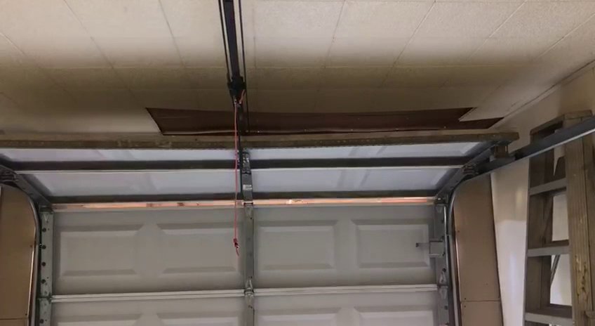 Interior view of a garage showing a white sectional door, opener rail, and damaged ceiling, indicating a repair job by OklaHome Overhead Doors in Tulsa, OK