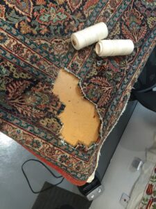 A severely damaged rug with a large hole, alongside spools of thread for repair at Renaissance Rug Cleaning Inc. in Portland, OR.