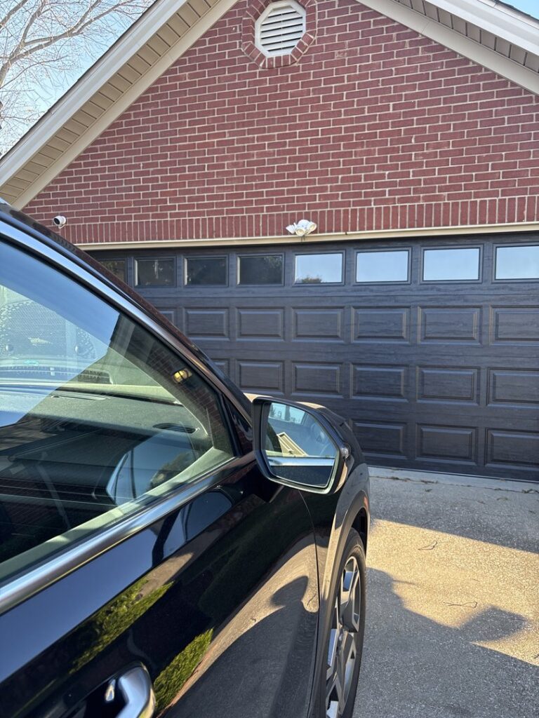 A dark grey garage door installed on a brick house, partially visible behind a car, by Precision Garage Door of Owensboro Evansville in Owensboro, KY.