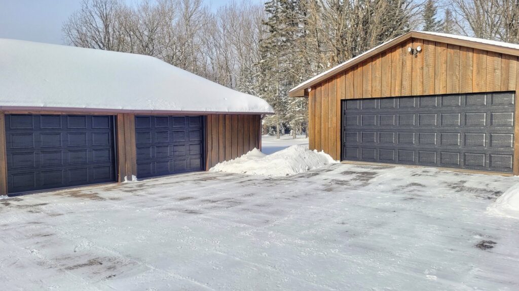 Two dark grey garage doors on wooden garages in a snowy winter setting by AJK Door Services in Duluth, MN.