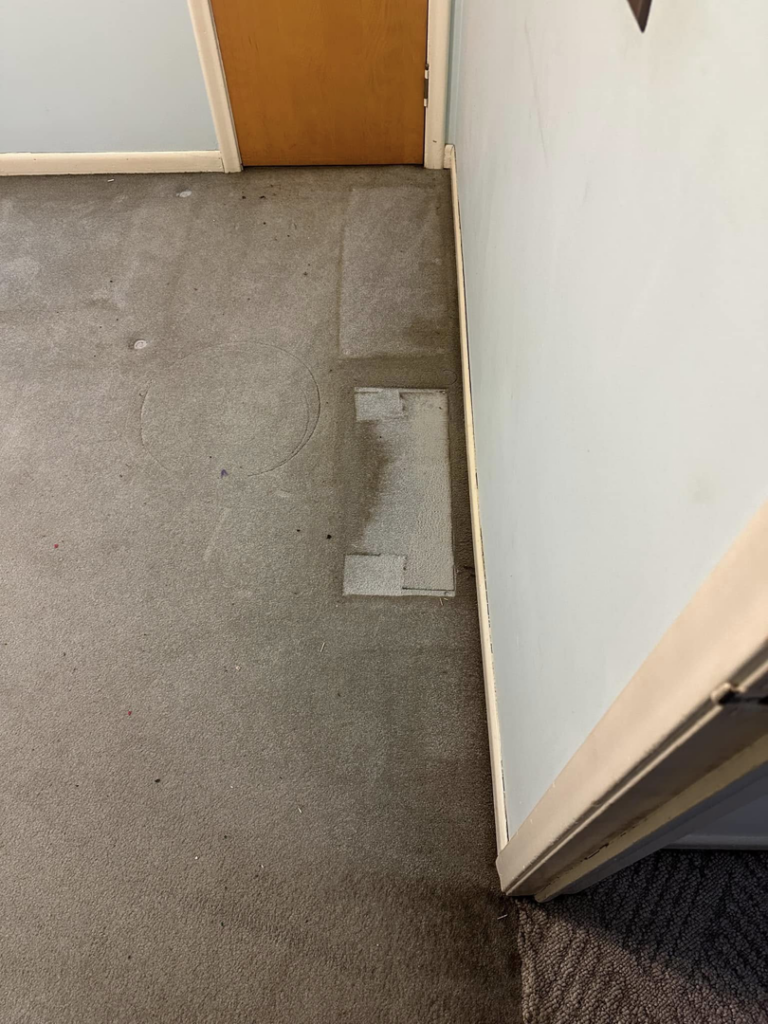 A dirty grey carpet with visible stains and a floor vent, awaiting professional cleaning by Triple B Carpet Cleaning in Roseville, MI.