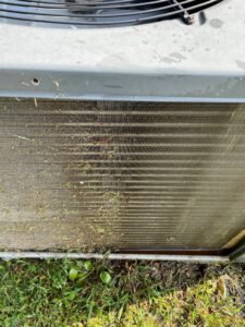 Close-up of a dirty outdoor HVAC condenser coil covered in grass and debris, ready for cleaning by Mission HVAC in Huntsville, AL.