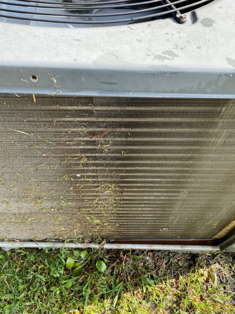 Close-up of a dirty outdoor HVAC condenser coil covered in grass and debris, ready for cleaning by Mission HVAC in Huntsville, AL.