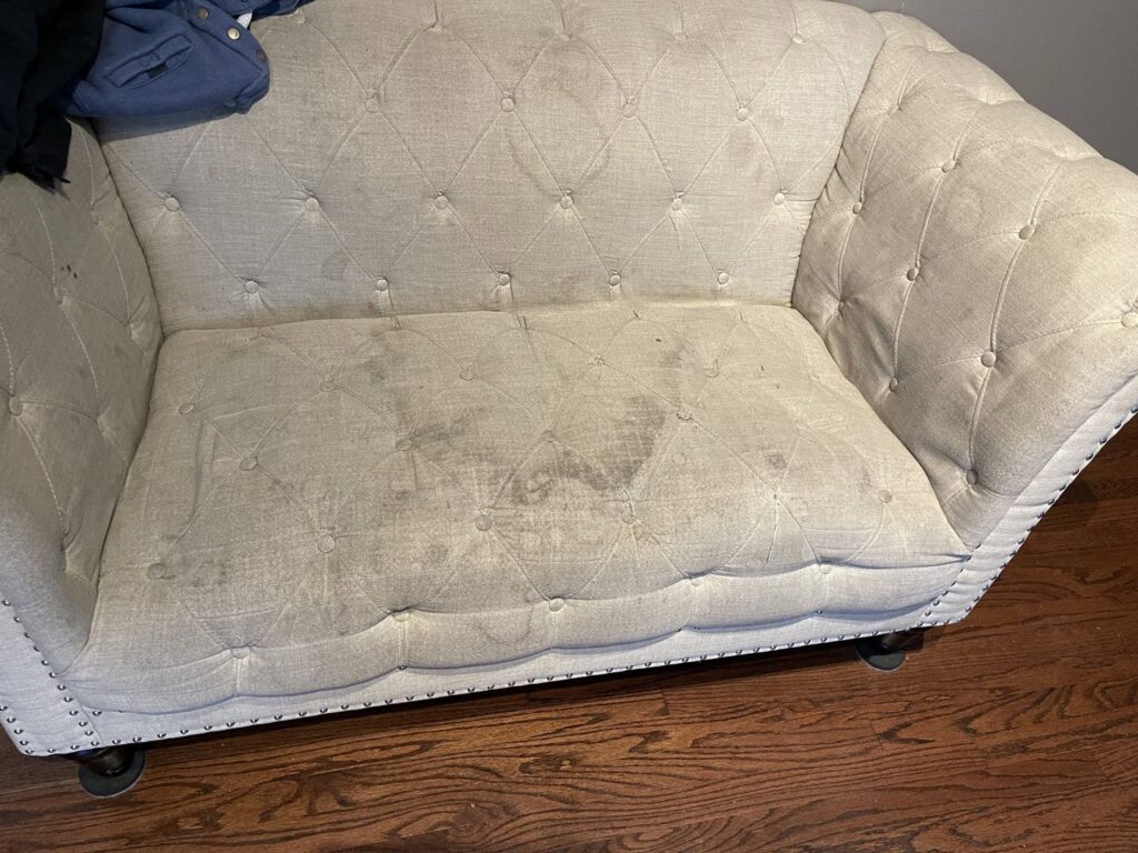 A dirty, light-colored upholstered loveseat with visible stains, indicating the need for professional cleaning by Steam Choice Carpet Care Inc in Chicago, IL.