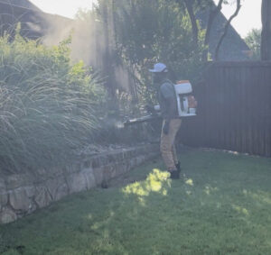 A Dr. Green Services employee applying pest control treatment with a backpack sprayer to shrubs and lawn edges in Joliet, IL