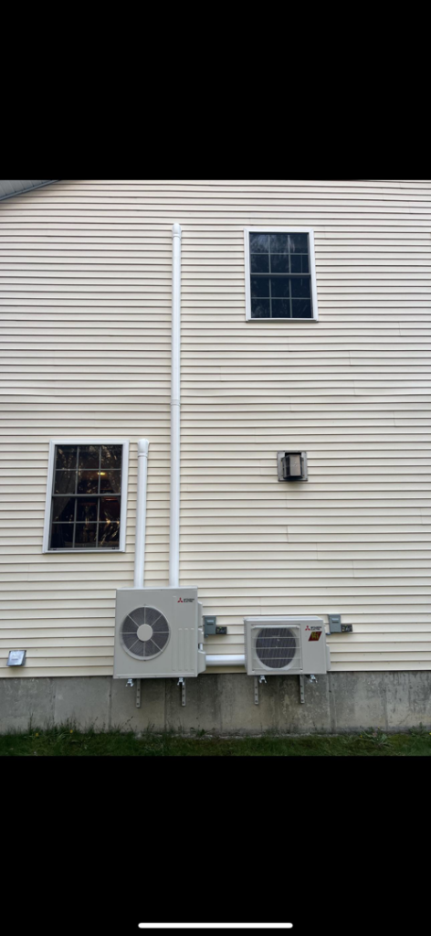 Two heat pump outdoor units installed on the yellow siding of a residential home by S&J Heat Pumps in Augusta, ME.