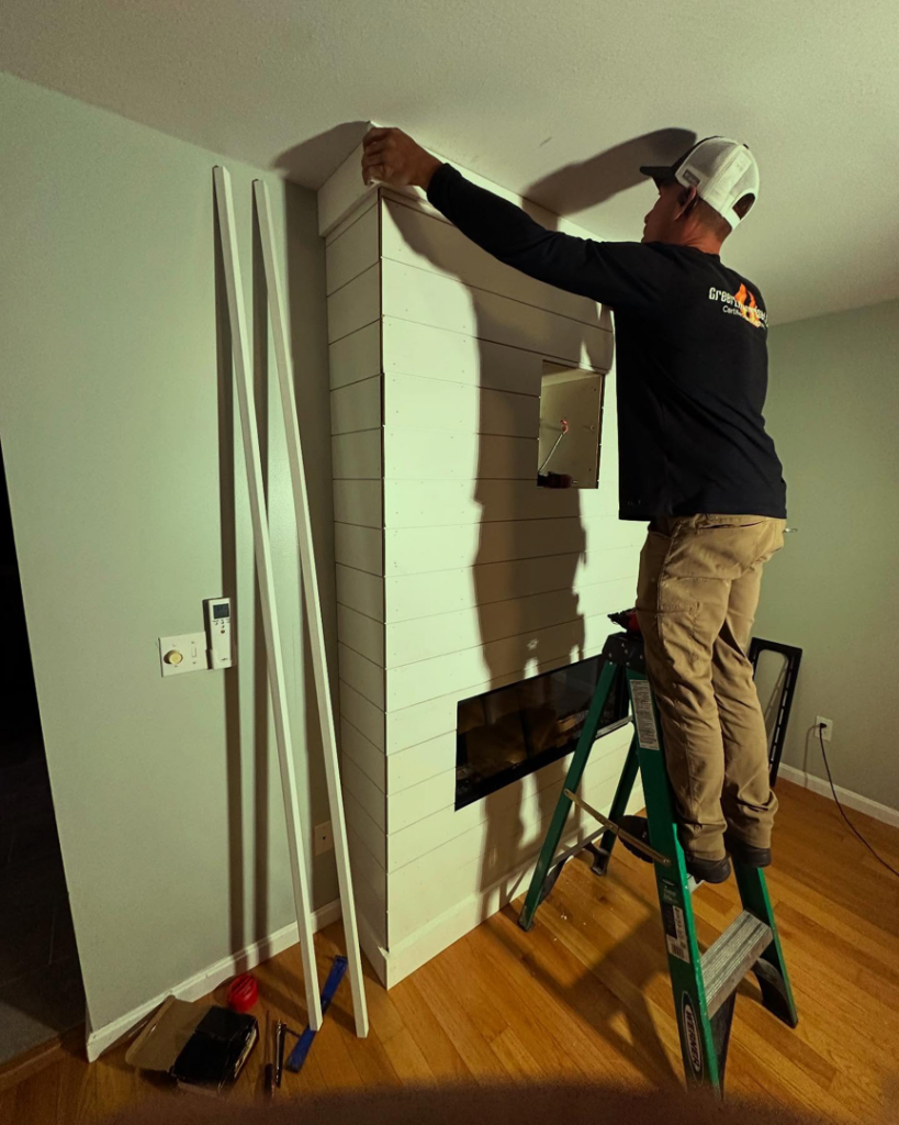Greer Enterprises technician on a ladder installing a custom electric fireplace surround in Raymond, NH.
