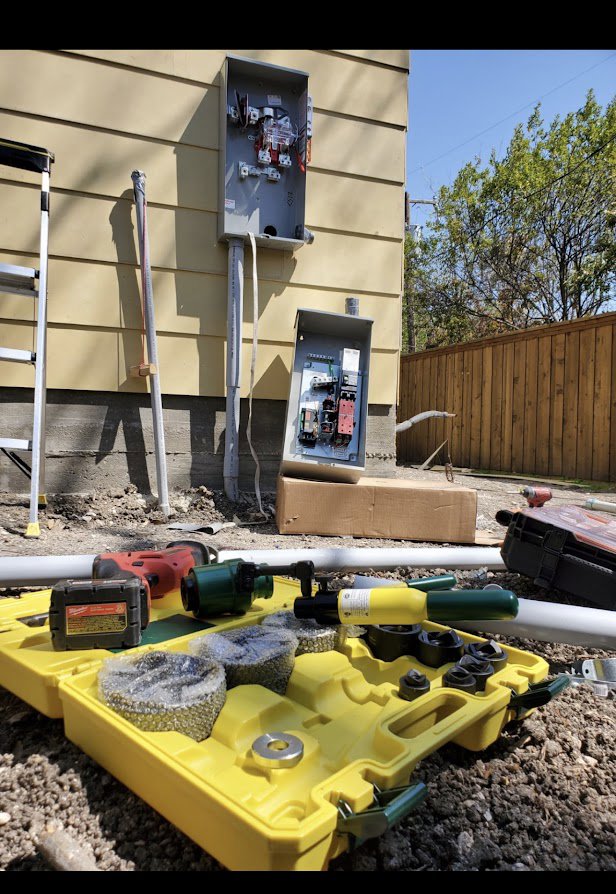 Electrical tools and conduit on the ground with an open electrical meter box on a building wall by Genesys Electrical Contractors in Dallas, TX