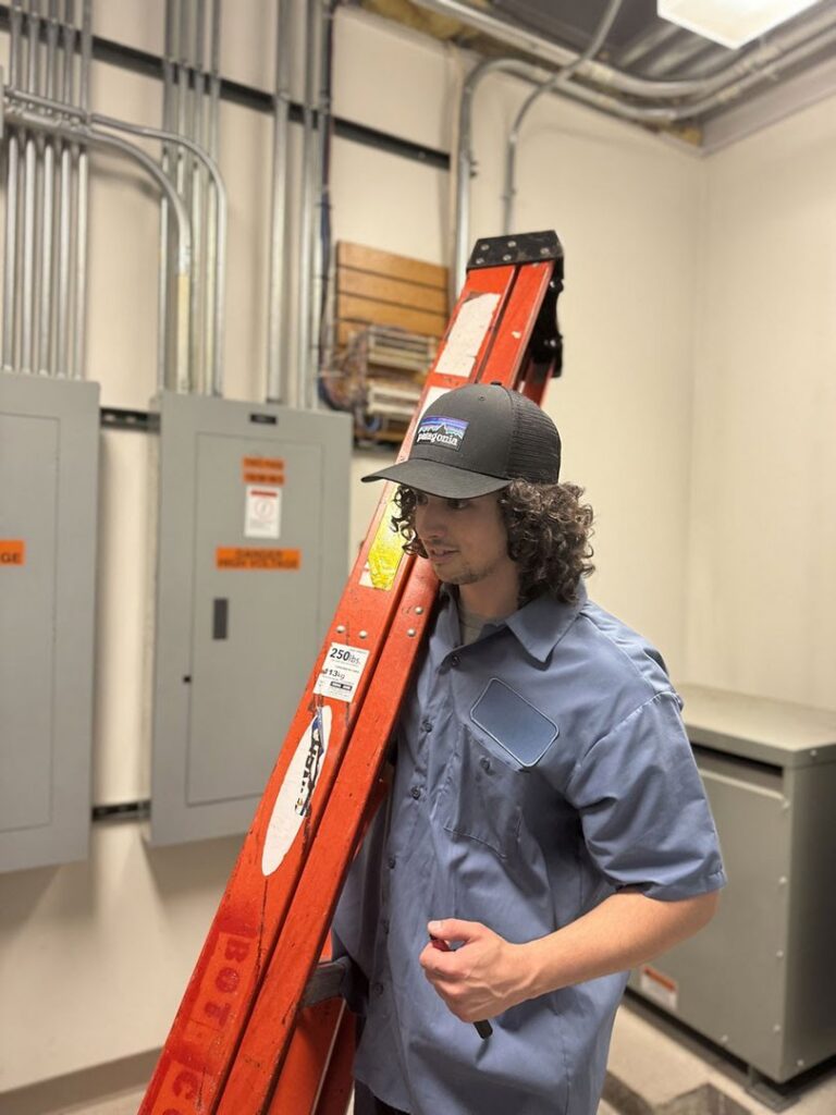 An electrician carrying a ladder in a room with electrical panels and conduits at Three Rivers Electric in Fort Wayne, IN.