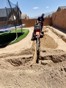 An electrician digging a trench in a backyard for underground electrical or data lines using a trenching machine by CAAM Integration LLC in Albuquerque, NM.