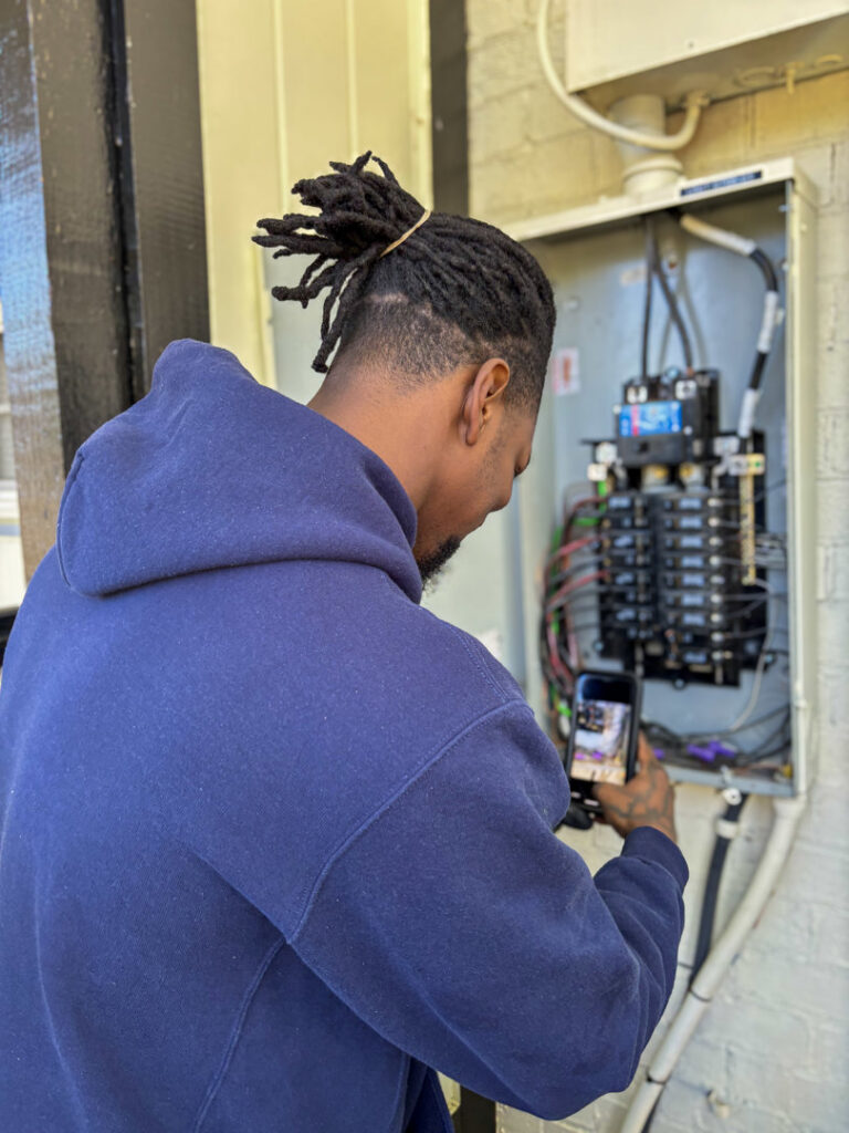 An electrician taking a photo of an open electrical panel with a smartphone, documenting work for Carolina Voltage in Charlotte, NC.