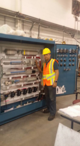 An electrician in a hard hat and safety vest inspecting a large industrial control panel for D-America Electrical Contractor LLC in Passaic, NJ.