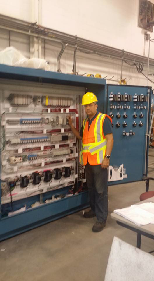 An electrician in a hard hat and safety vest inspecting a large industrial control panel for D-America Electrical Contractor LLC in Passaic, NJ.