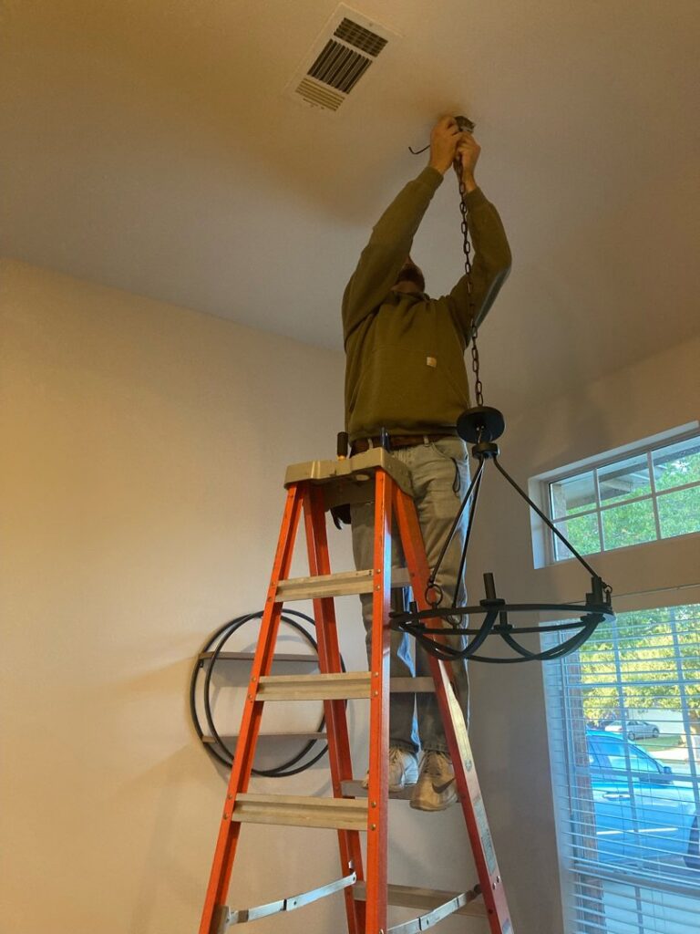 An electrician on a ladder installing a ceiling chandelier, showcasing professional lighting installation by Volta Electrical LLC in Plano, TX.