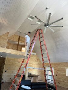 An electrician on a tall ladder installing a large ceiling fan in a high-ceiling room, performing work for Greif Electric Company in Dolgeville, NY.