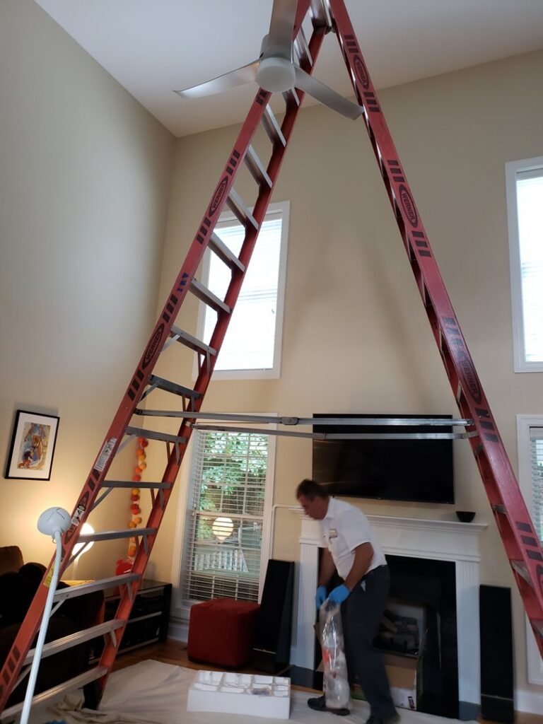 An electrician from IRV Plumbing, Electric & HVAC on a ladder installing a ceiling fan in a home in Charlotte, NC