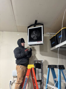 An electrician installing a ceiling-mounted heater or fan unit near an electrical panel for LeBrun Electric, Heating & Cooling in Brooklyn Park, MN.