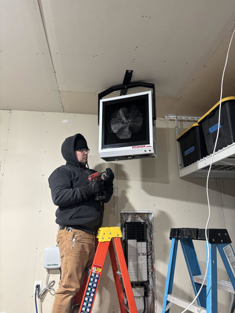 An electrician installing a ceiling-mounted heater or fan unit near an electrical panel for LeBrun Electric, Heating & Cooling in Brooklyn Park, MN.