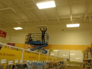 An electrician from Sabteck Electric installing a large ceiling light fixture from a scissor lift inside a commercial building in Timmins, ON